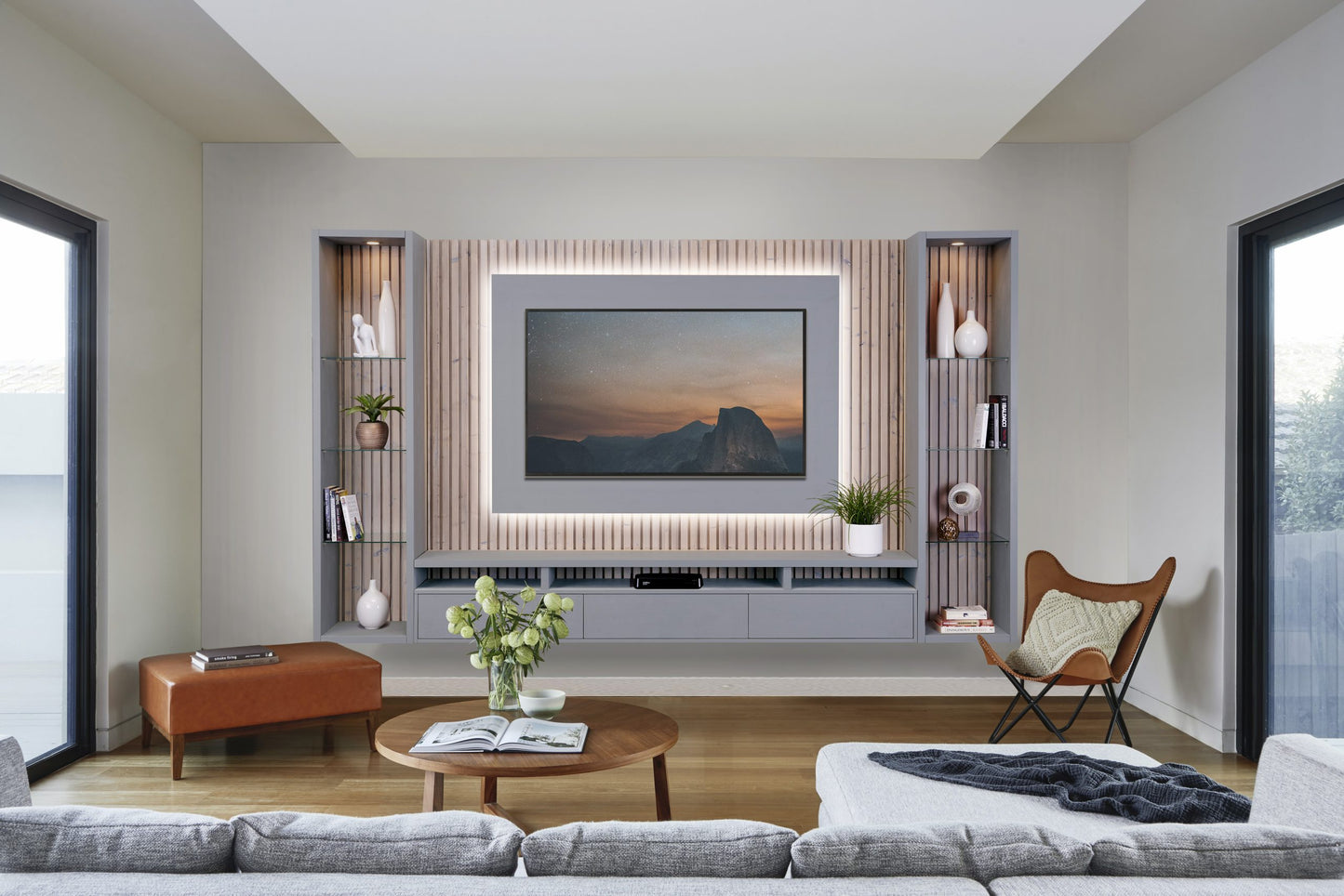 A minimalist living room featuring a large, mounted television surrounded by blush-toned paneling, flanked by symmetrical floating shelving with decorative items, a cozy grey sofa, a chic leather ottoman